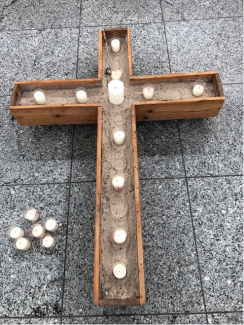 Ein Kreuz aus Holz und Sand liegt auf dem Boden, darauf sind Kerzen abgestellt